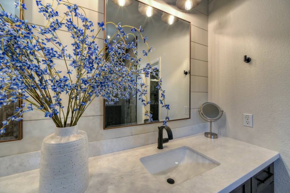 A bathroom with a sink, mirror and flowers in the corner.